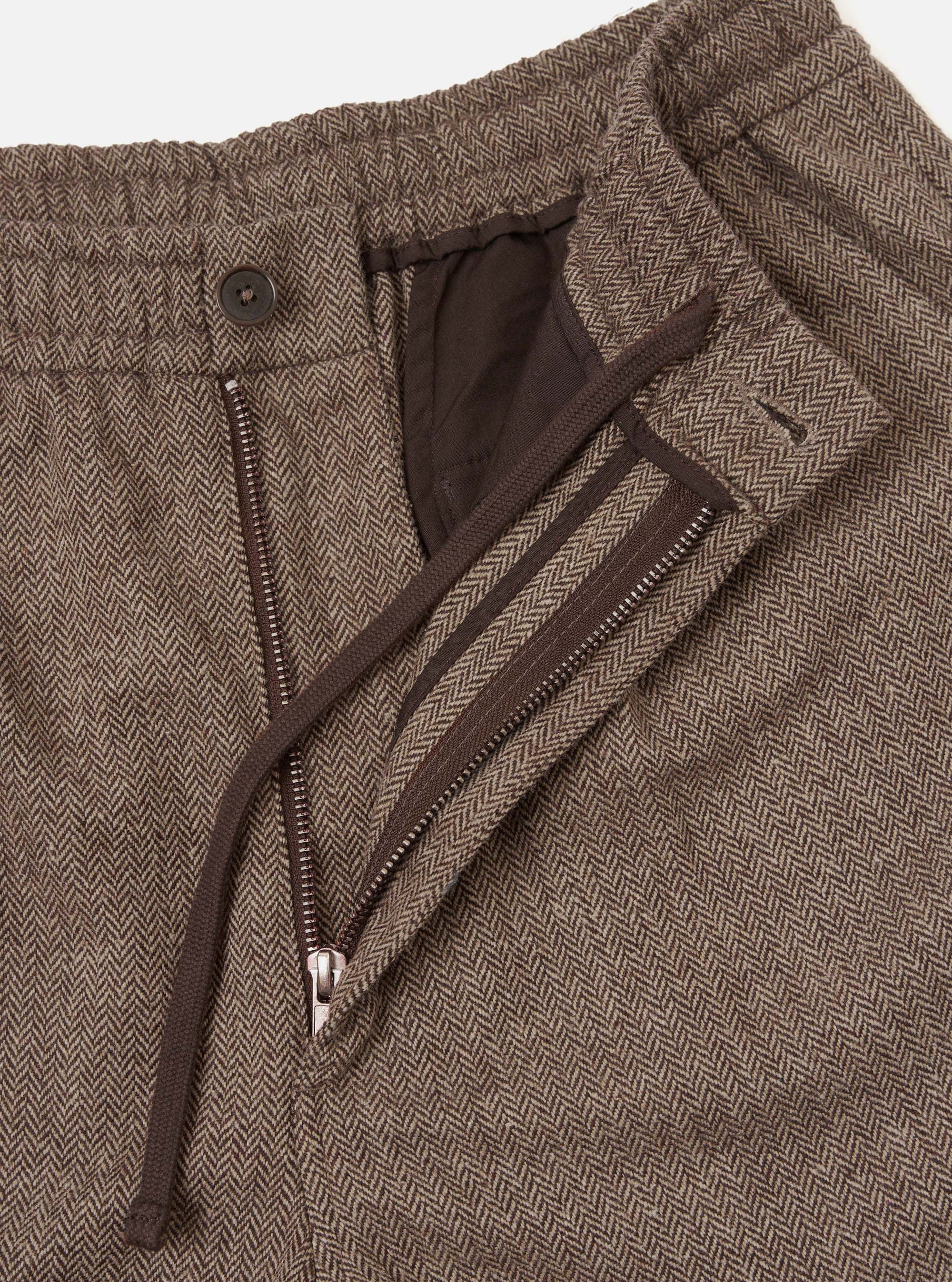 Close-up detail shot of the brown wool herringbone Gillespie pant on a white studio backdrop. The fly is open, showing a metal zip, a dark brown button at the waistband, a zip-fastened welt pocket on the fly flap, and a dark brown lining. The herringbone weave is clearly visible across the fabric.
