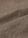 Close-up detail shot of the brown wool herringbone fabric used in the Gillespie Pant. The weave shows a distinct V-shaped herringbone pattern in dark brown and ecru tones. The fabric is folded to reveal texture and weight, with a slightly flecked, woollen surface visible across the folds.