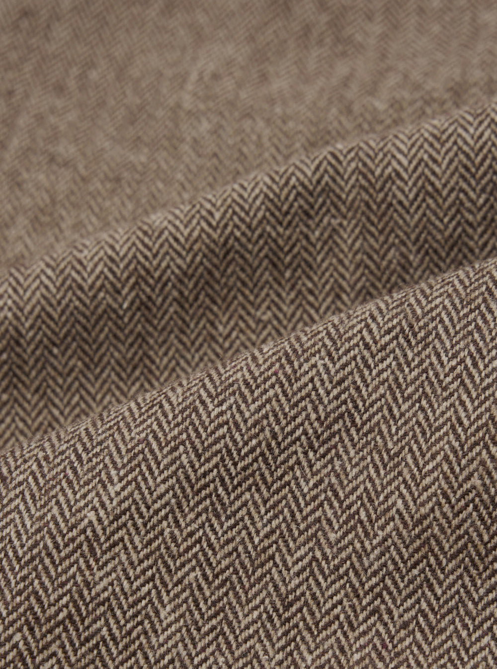 Close-up detail shot of the brown wool herringbone fabric used in the Gillespie Pant. The weave shows a distinct V-shaped herringbone pattern in dark brown and ecru tones. The fabric is folded to reveal texture and weight, with a slightly flecked, woollen surface visible across the folds.