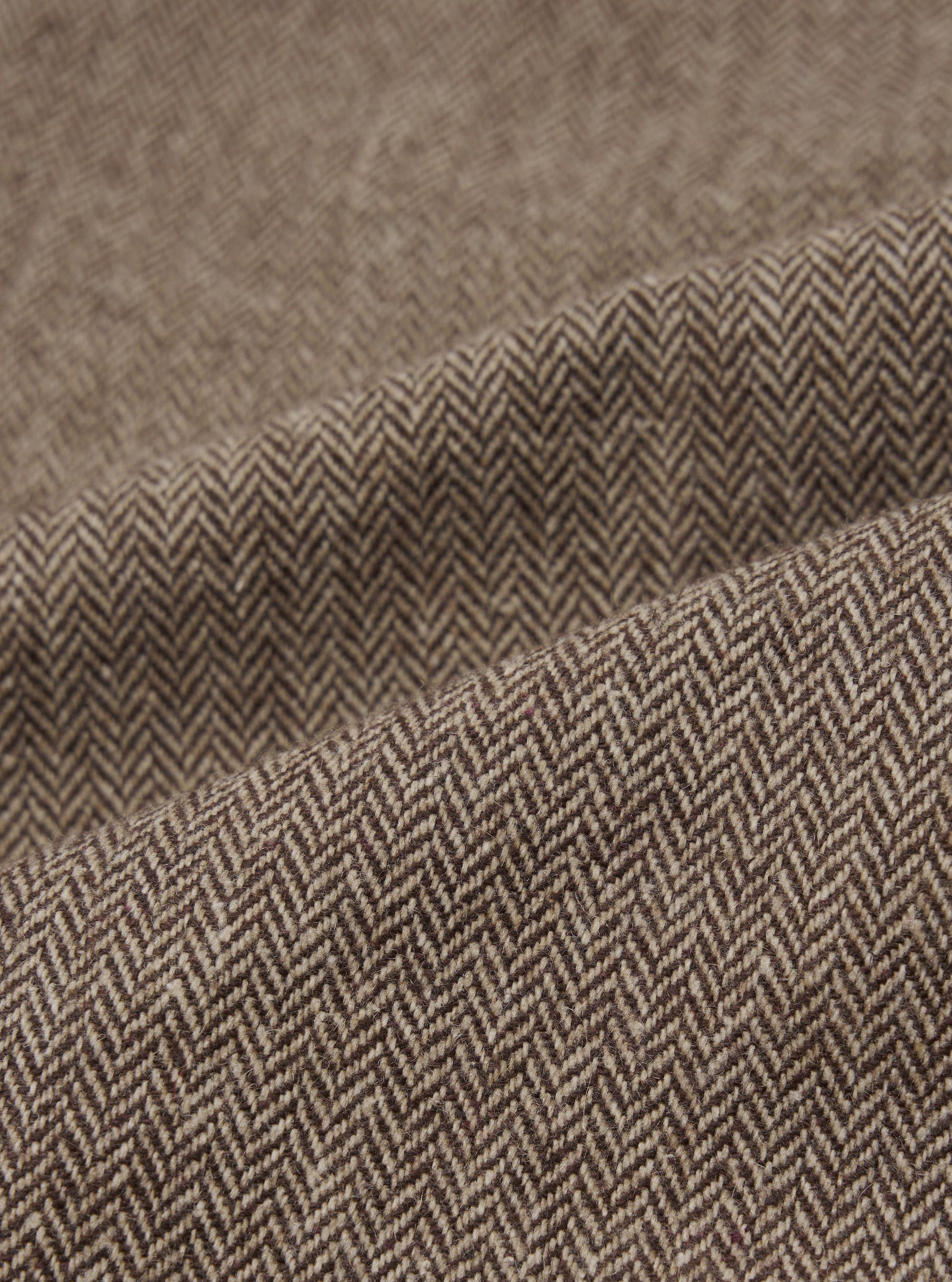 Close-up detail shot of the brown wool herringbone fabric used in the Gillespie Pant. The weave shows a distinct V-shaped herringbone pattern in dark brown and ecru tones. The fabric is folded to reveal texture and weight, with a slightly flecked, woollen surface visible across the folds.