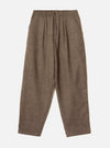 Brown wool herringbone pleated trousers laid flat on a light grey studio backdrop. The fabric shows a fine herringbone weave throughout. A structured waistband features a single button fastening and slanted side pockets. Double pleats run from the waistband to full-length tapered legs with plain hems.