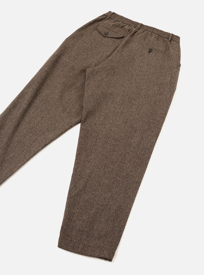 Brown wool herringbone track pants laid flat on a white studio surface, angled diagonally. The herringbone weave is clearly visible across the tapered leg. The elasticated waistband features a buttoned flap pocket at the rear left and a small button-fastened coin pocket. A single pleat runs down the front of each leg.