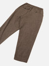 Brown wool herringbone track pants laid flat on a white studio surface, angled diagonally. The herringbone weave is clearly visible across the tapered leg. The elasticated waistband features a buttoned flap pocket at the rear left and a small button-fastened coin pocket. A single pleat runs down the front of each leg.