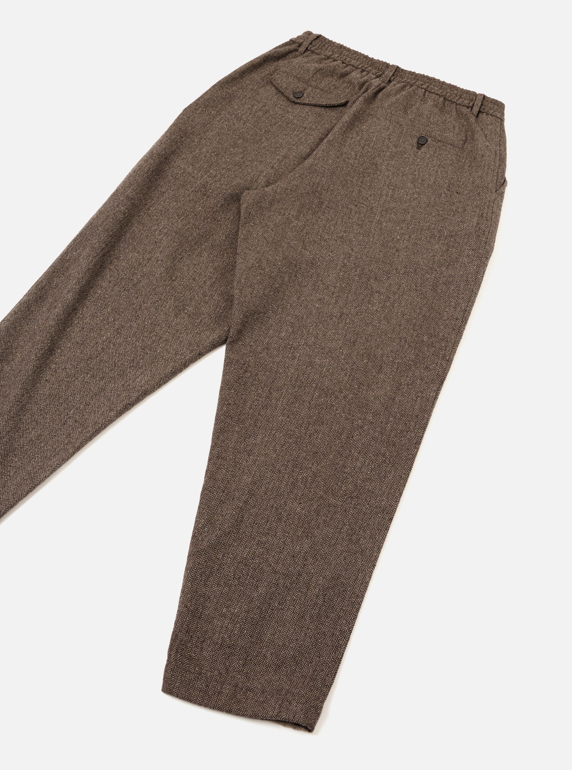 Brown wool herringbone track pants laid flat on a white studio surface, angled diagonally. The herringbone weave is clearly visible across the tapered leg. The elasticated waistband features a buttoned flap pocket at the rear left and a small button-fastened coin pocket. A single pleat runs down the front of each leg.