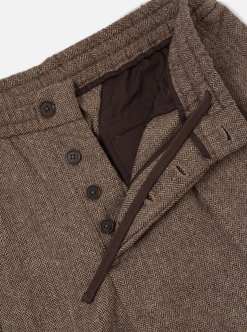 Close-up flat lay of brown wool herringbone pleated track pants on a light grey backdrop. The waistband is open, revealing a dark brown drawcord and inner lining. Four dark horn buttons fasten the fly placket. The herringbone weave texture is prominent throughout.