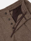 Close-up flat lay of brown wool herringbone pleated track pants on a light grey backdrop. The waistband is open, revealing a dark brown drawcord and inner lining. Four dark horn buttons fasten the fly placket. The herringbone weave texture is prominent throughout.
