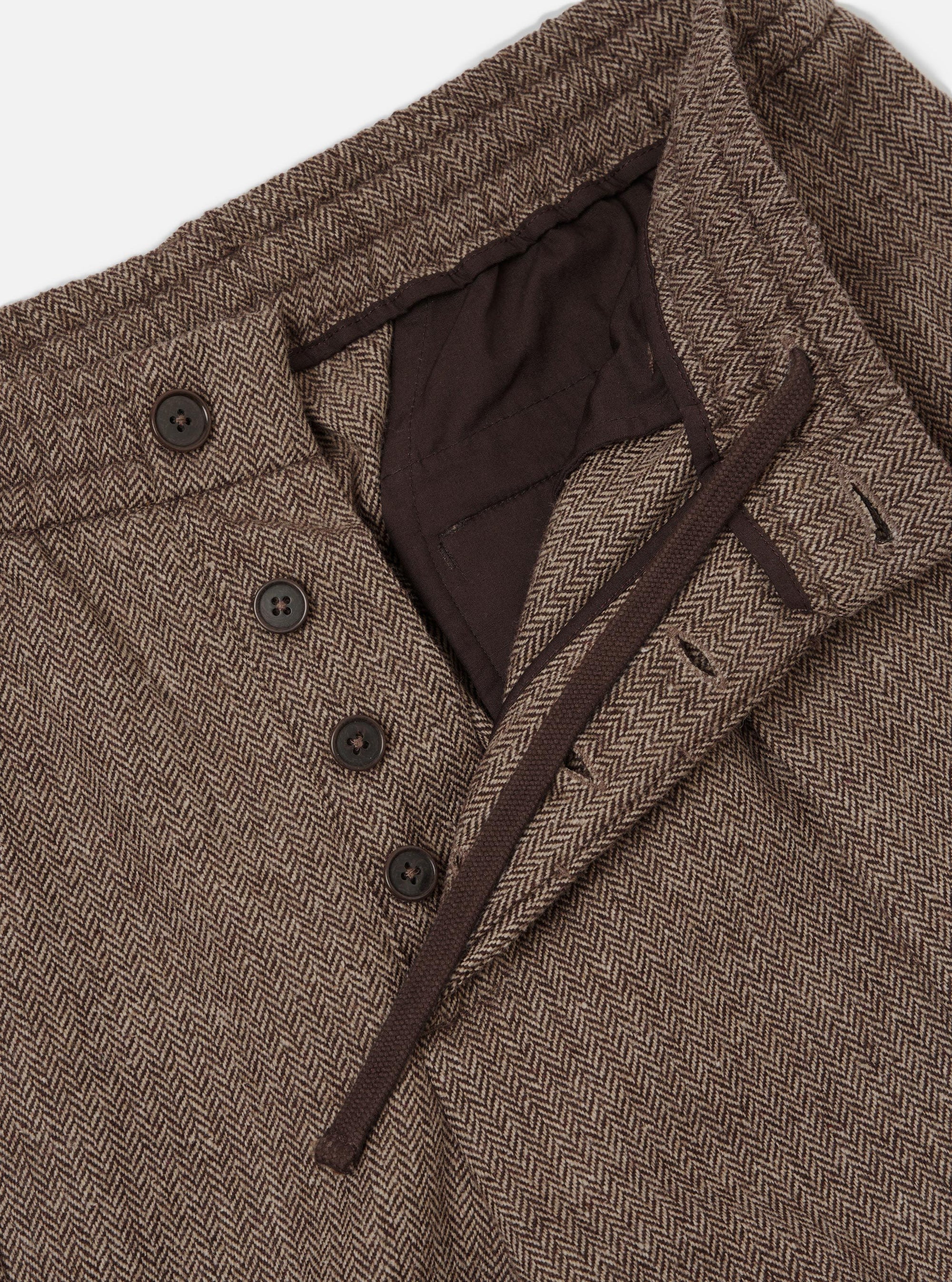Close-up flat lay of brown wool herringbone pleated track pants on a light grey backdrop. The waistband is open, revealing a dark brown drawcord and inner lining. Four dark horn buttons fasten the fly placket. The herringbone weave texture is prominent throughout.