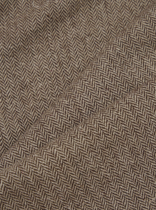Close-up detail shot of brown wool herringbone fabric. The tightly woven chevron pattern is visible in dark brown and ecru tones across folded and creased sections of the trouser cloth, showing the texture and weight of the material.
