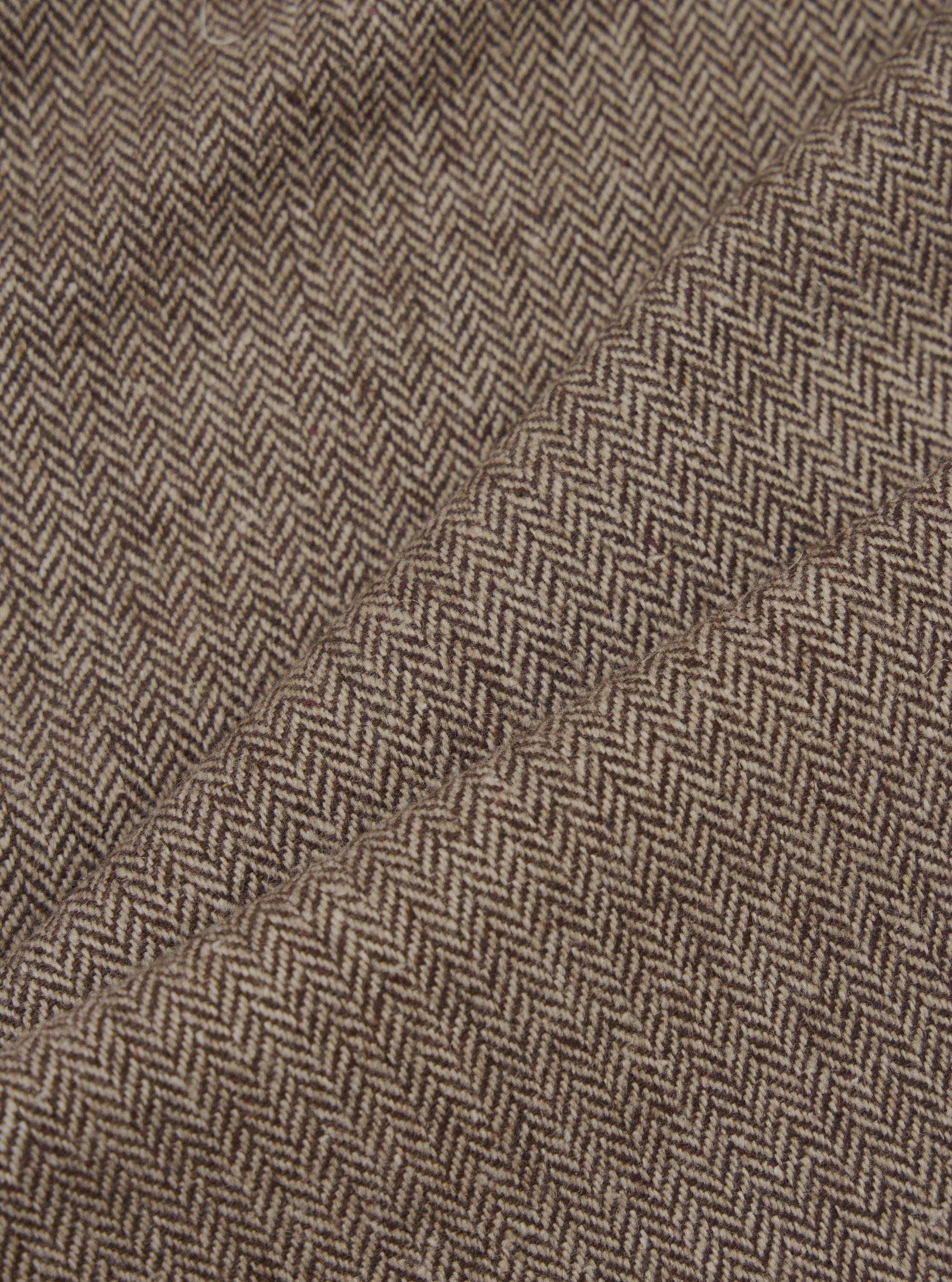 Close-up detail shot of brown wool herringbone fabric. The tightly woven chevron pattern is visible in dark brown and ecru tones across folded and creased sections of the trouser cloth, showing the texture and weight of the material.