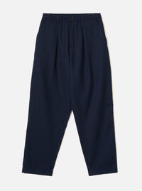 Navy wool herringbone pleated track pants laid flat on a light grey studio backdrop. The elasticated waistband features a single dark metal button at the centre front. Two slant side pockets are visible. A single pleat runs down each leg. The trousers have a relaxed, tapered fit with a cropped hem.