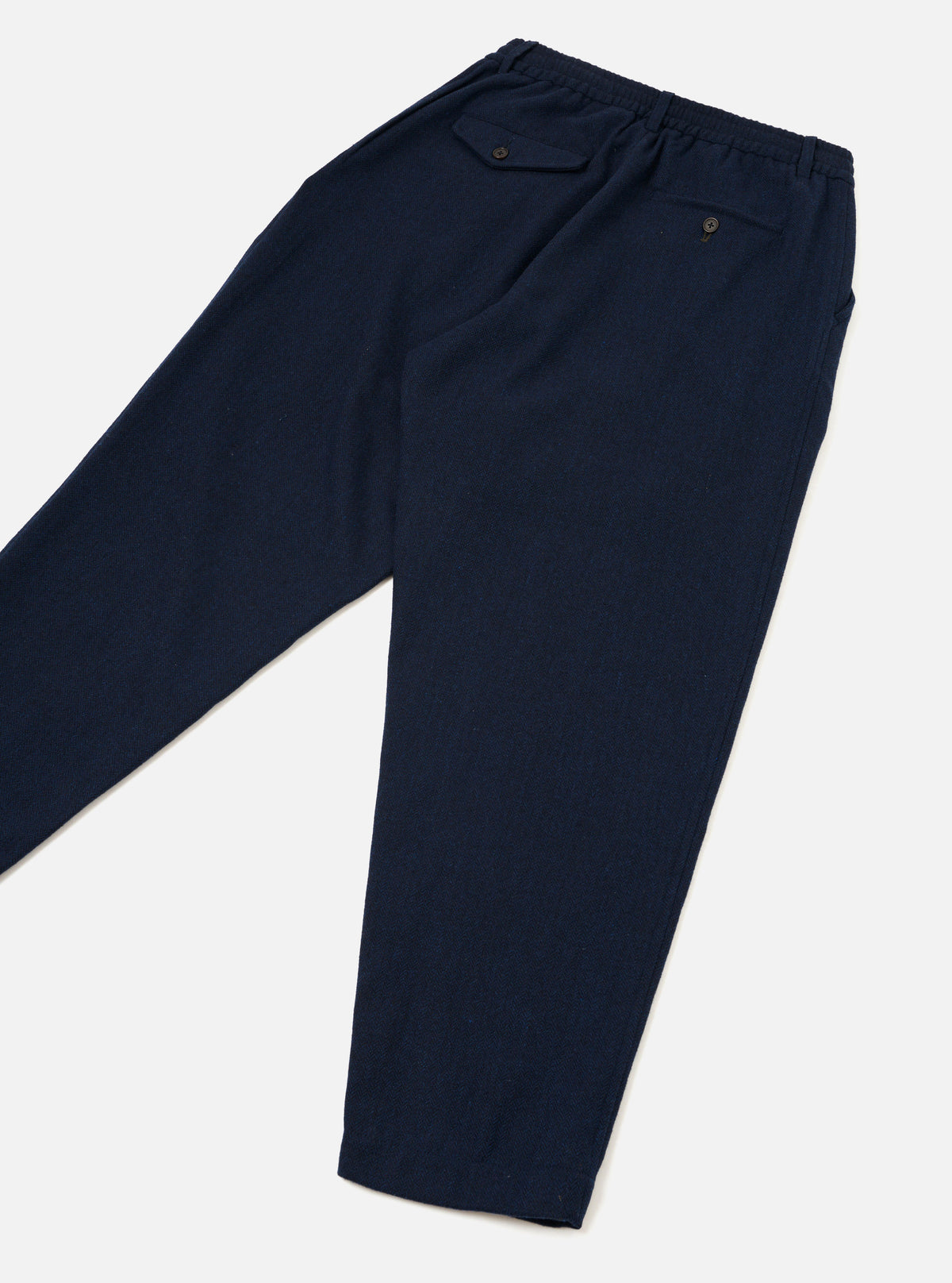Navy wool herringbone track pants laid flat on a light grey surface, showing the back. An elasticated waistband sits at the top with two button-fastened welt pockets visible. The fabric has a fine herringbone texture. The tapered leg narrows toward the plain, unfinished hem.