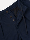 Close-up of the waistband and fly area of navy wool herringbone pleated track pants. Three dark horn buttons fasten the fly flap, with a visible black lining behind the open waistband. A drawcord sits inside the waistband. The herringbone weave texture is clearly visible. Plain white studio backdrop.