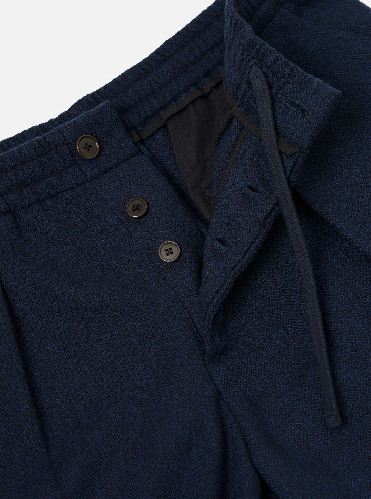 Close-up of the waistband and fly area of navy wool herringbone pleated track pants. Three dark horn buttons fasten the fly flap, with a visible black lining behind the open waistband. A drawcord sits inside the waistband. The herringbone weave texture is clearly visible. Plain white studio backdrop.