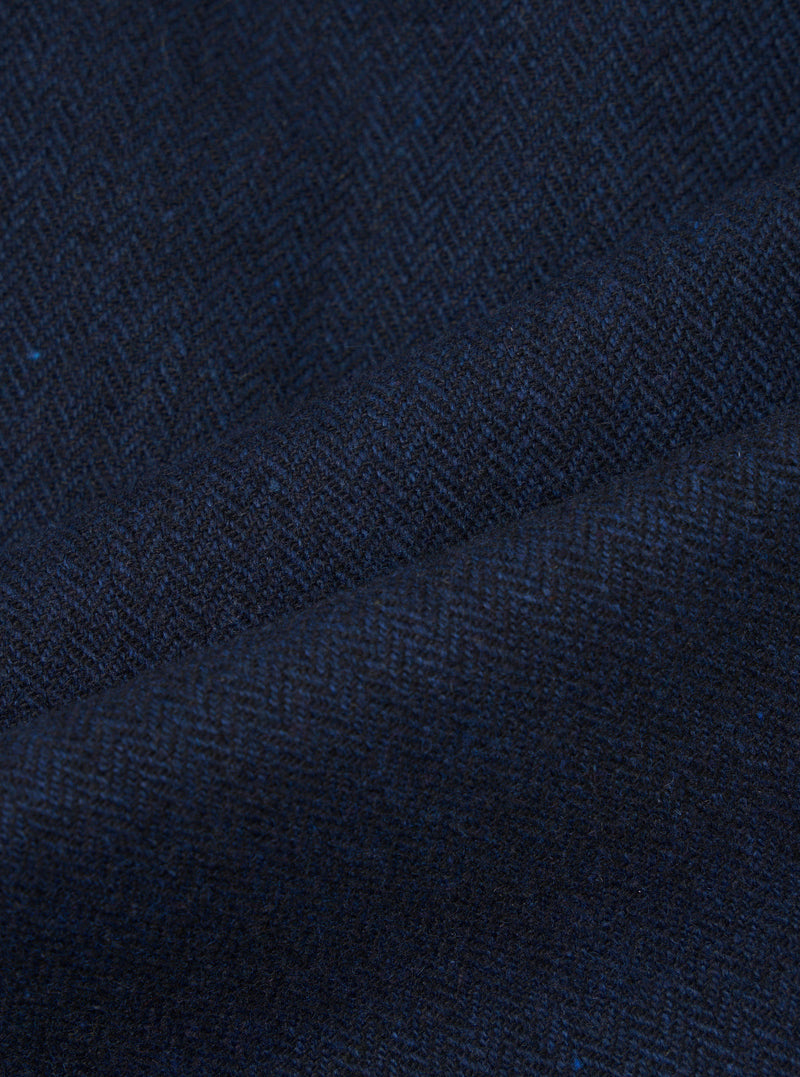 Close-up of navy wool herringbone fabric, showing the diagonal zigzag weave pattern in deep navy and dark charcoal tones with flecks of bright blue. The cloth is gathered and folded, highlighting the texture and weight of the woven wool.