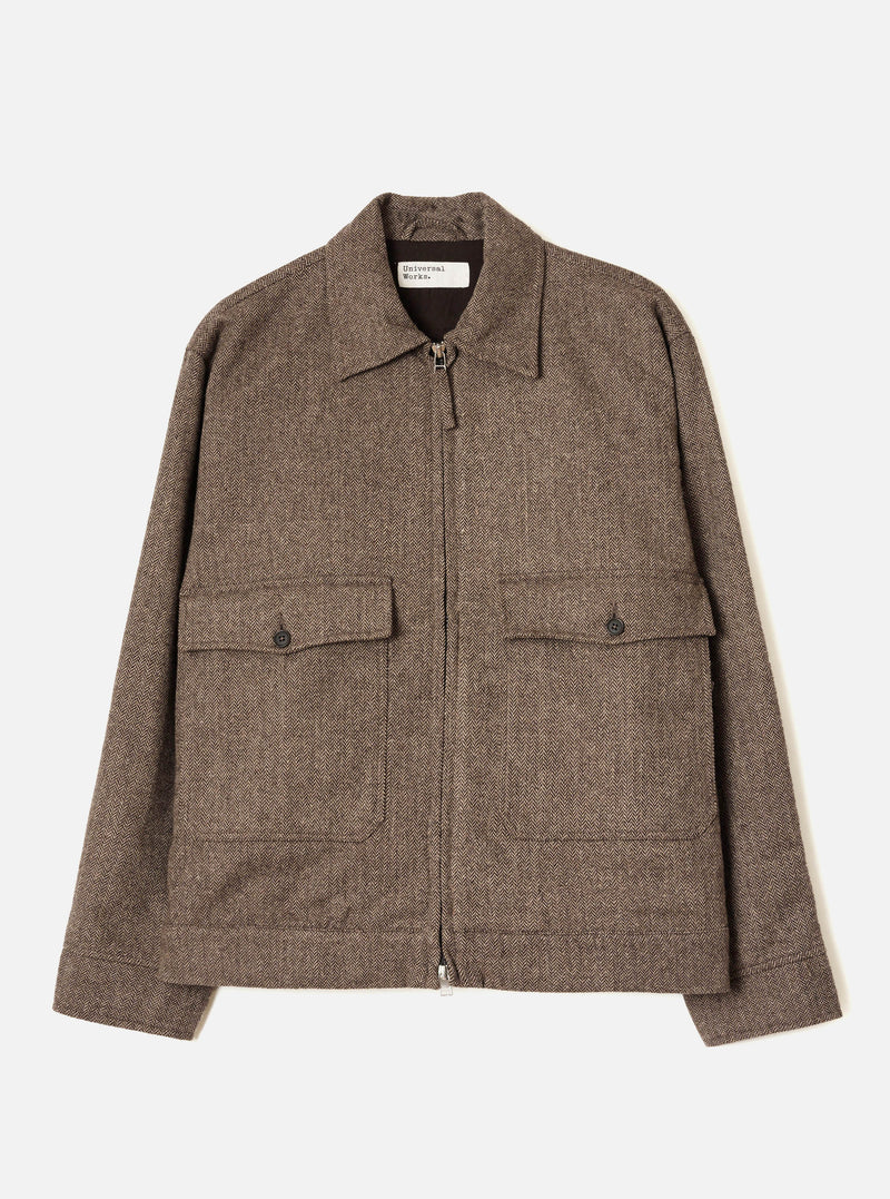 Brown wool herringbone zip-through jacket, flat lay on a plain light grey background. Features a point collar, central metal zip, two large chest patch pockets with button-fastened flaps, and plain cuffs. A Universal Works label is visible at the neck. The dark brown lining is visible at the collar.