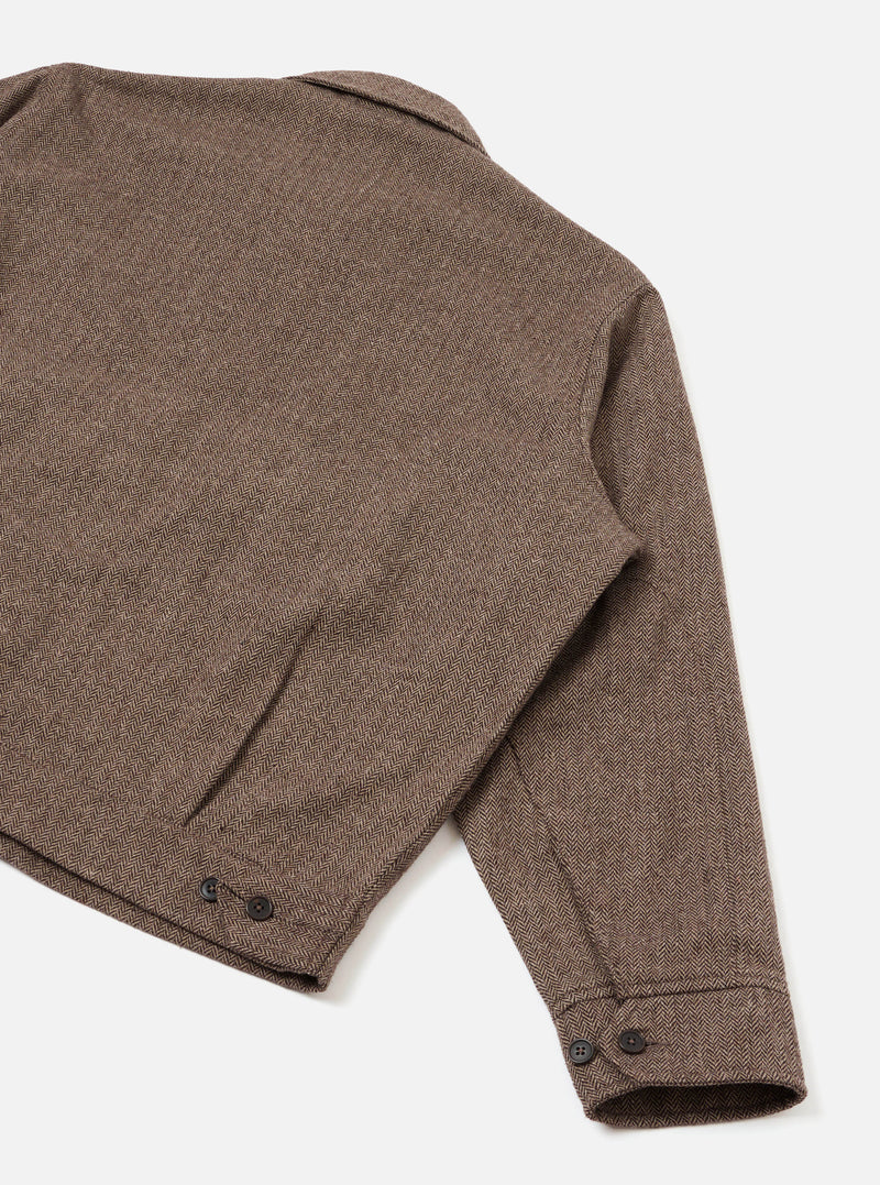 Close-up of the rear and sleeve of a brown wool herringbone jacket on a white studio backdrop. The herringbone weave is clearly visible across the back panel and long sleeve. A welt pocket sits at the lower back. Dark buttons fasten the cuff and appear at the hem. No model or mannequin is present.