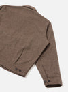 Close-up of the rear and sleeve of a brown wool herringbone jacket on a white studio backdrop. The herringbone weave is clearly visible across the back panel and long sleeve. A welt pocket sits at the lower back. Dark buttons fasten the cuff and appear at the hem. No model or mannequin is present.