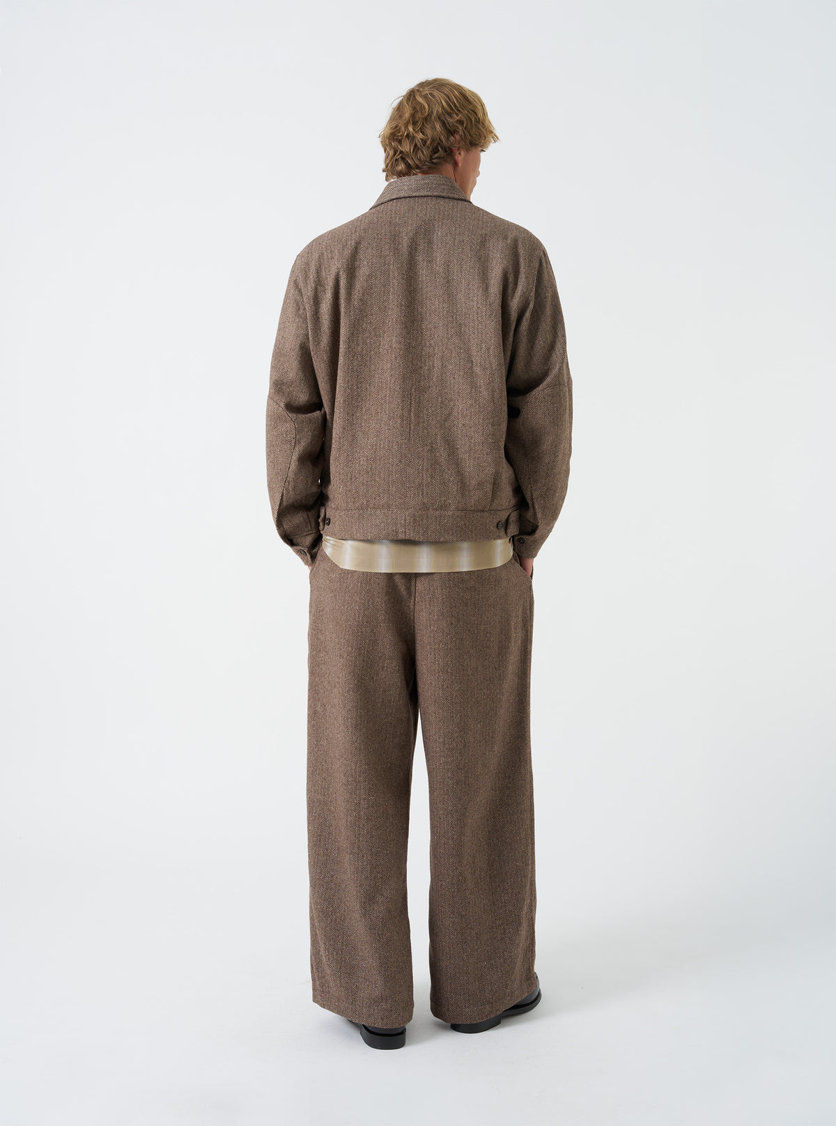 A back view of a model wearing the Brown Wool Herringbone E130 Jacket. The image highlights the rear construction of the brown wool-blend coach jacket, featuring a clean shoulder line and straight hem set against a plain backdrop.