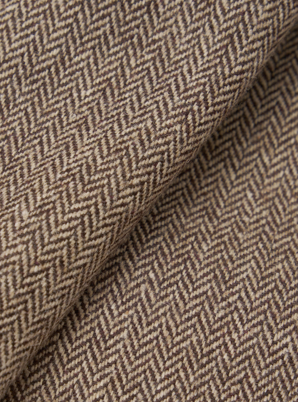 Close-up detail shot of a brown wool herringbone jacket fabric. The weave shows a classic chevron pattern in dark brown and oatmeal, with a lightly textured, slightly hairy wool surface. The fabric is folded to reveal depth and drape. No background is visible.