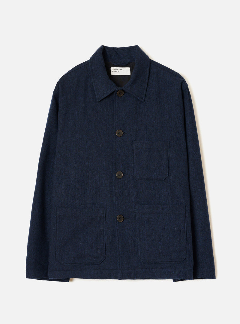 Navy wool herringbone chore jacket laid flat on a light grey studio backdrop. Features a point collar, four dark horn buttons down the centre front, one chest patch pocket and two large lower patch pockets. Full-length sleeves with plain cuffs. A Universal Works woven label sits at the back neck. Subtle herringbone weave texture visible throughout.