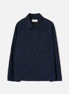 Navy wool herringbone chore jacket laid flat on a light grey studio backdrop. Features a point collar, four dark horn buttons down the centre front, one chest patch pocket and two large lower patch pockets. Full-length sleeves with plain cuffs. A Universal Works woven label sits at the back neck. Subtle herringbone weave texture visible throughout.