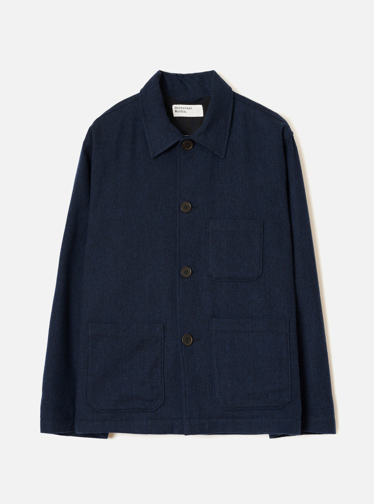 Navy wool herringbone chore jacket laid flat on a light grey studio backdrop. Features a point collar, four dark horn buttons down the centre front, one chest patch pocket and two large lower patch pockets. Full-length sleeves with plain cuffs. A Universal Works woven label sits at the back neck. Subtle herringbone weave texture visible throughout.