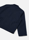Flat lay of a navy wool herringbone Ives jacket on a plain white background, shot from above at an angle. The back and right sleeve are prominent, showing the fine herringbone texture of the fabric. A small epaulette sits at the shoulder seam, and a welt pocket is visible at the lower left. The plain cuff and straight hem are visible.