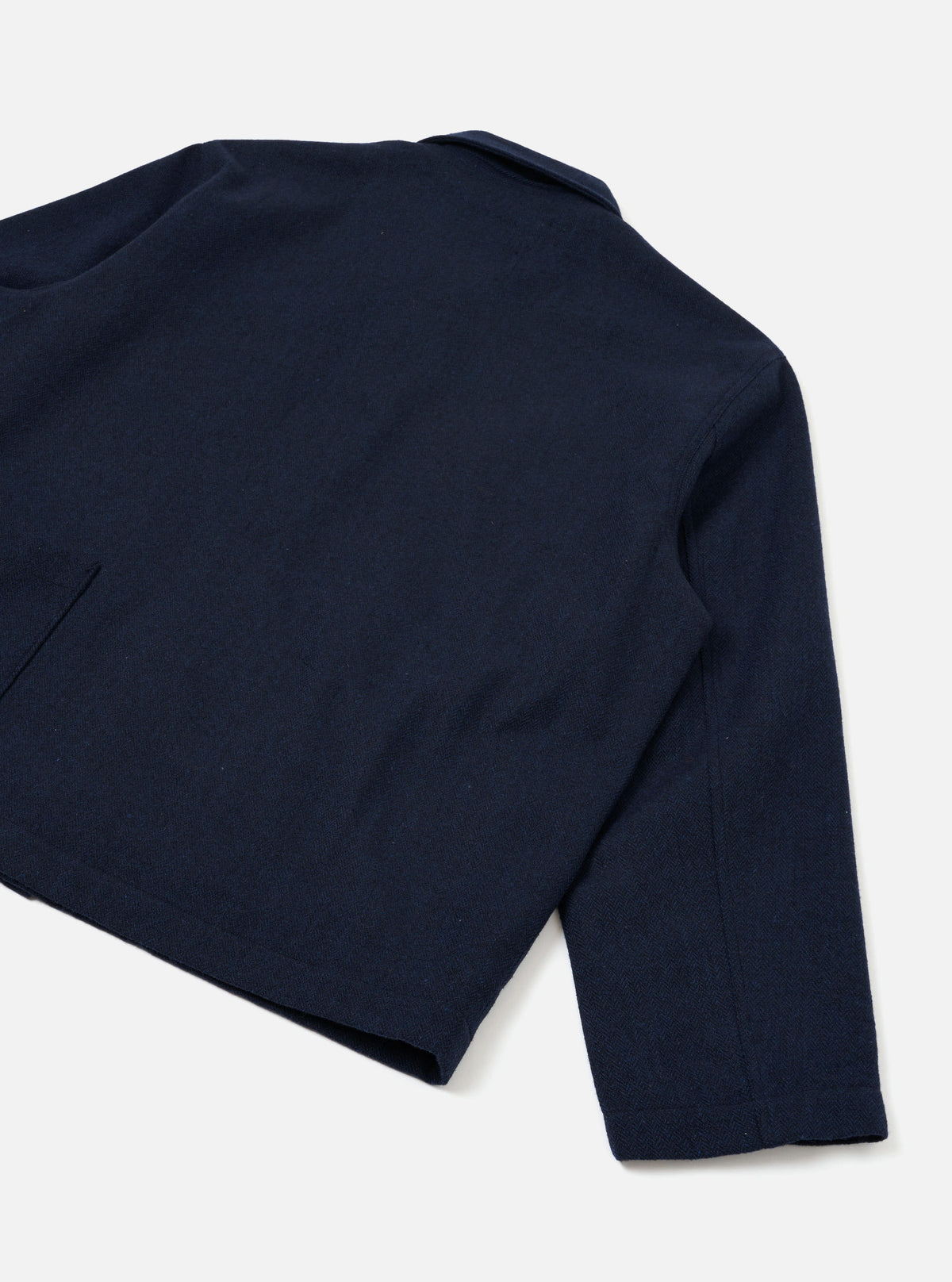 Flat lay of a navy wool herringbone Ives jacket on a plain white background, shot from above at an angle. The back and right sleeve are prominent, showing the fine herringbone texture of the fabric. A small epaulette sits at the shoulder seam, and a welt pocket is visible at the lower left. The plain cuff and straight hem are visible.