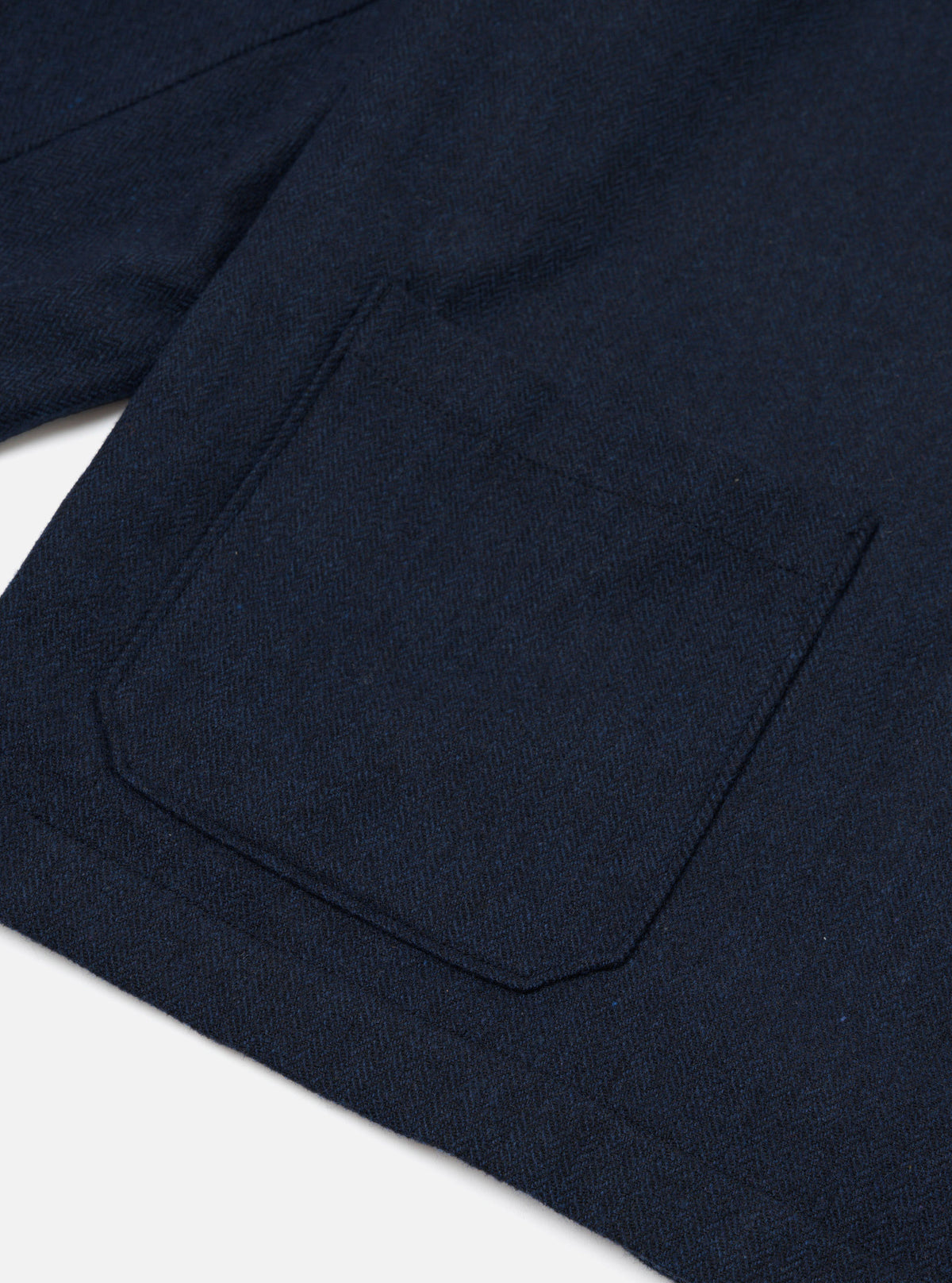 Close-up detail of the navy wool herringbone Ives Jacket laid flat on a white surface, showing a large rounded patch pocket and partial view of a second pocket above. The herringbone weave is clearly visible across the dense, textured fabric.