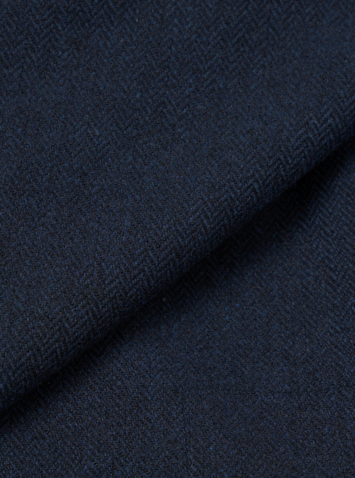 Close-up of navy wool herringbone fabric from the Ives Jacket, folded to show texture and weight. The tight herringbone weave shows dark navy and charcoal tones throughout. No background visible, the shot fills the frame with fabric detail only.