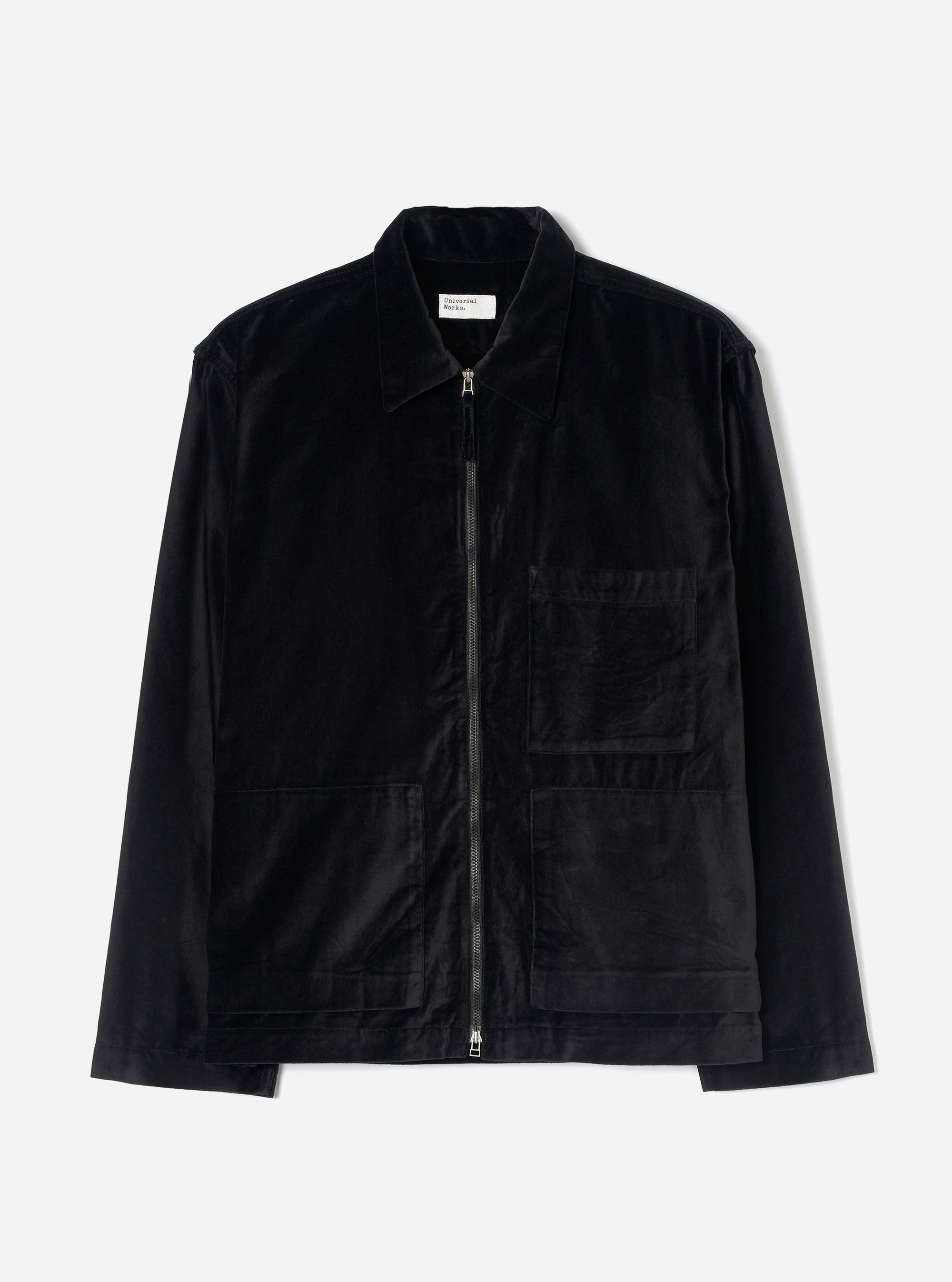 Black velvet zip-through overshirt on a plain light grey studio backdrop. Features a point collar, full-length black zip with silver metal pull tabs at top and bottom, one chest patch pocket, two lower patch pockets, and long plain sleeves. A woven Universal Works label sits inside the collar.