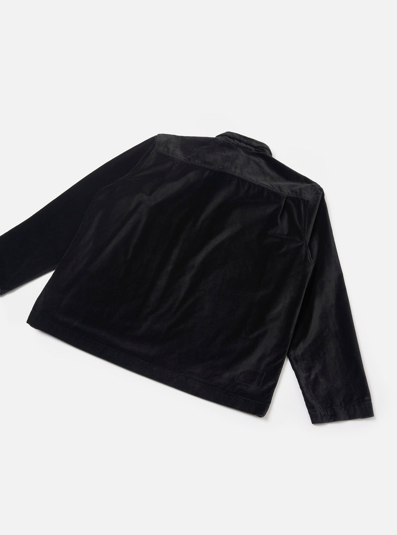 Black velvet zip overshirt laid flat on a light grey seamless backdrop, shot from a rear three-quarter angle. The soft, lustrous velvet fabric catches light across the back panel and long sleeves. A yoke seam runs across the upper back. A small woven label is visible at the lower left hem.