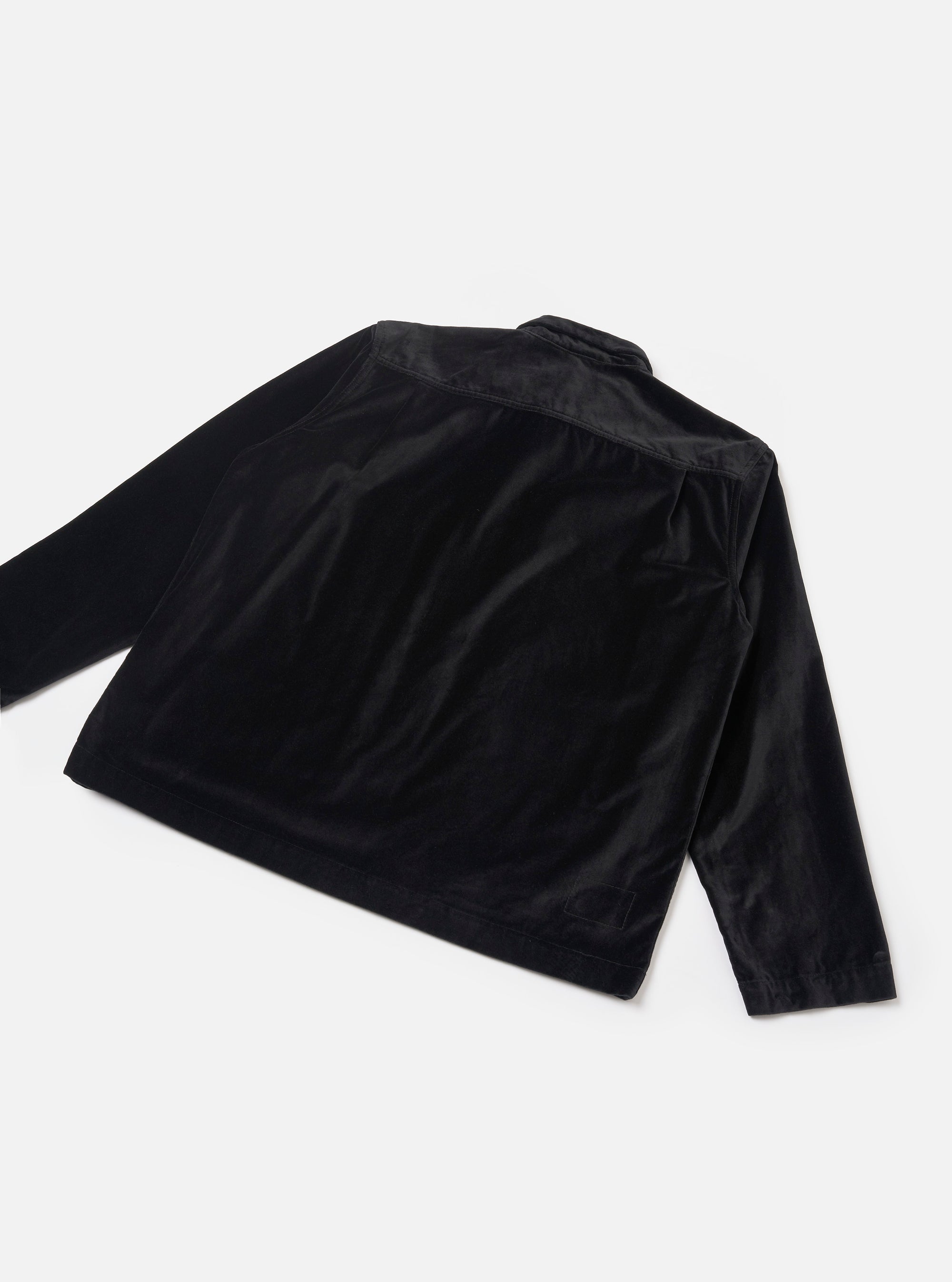 Black velvet zip overshirt laid flat on a light grey seamless backdrop, shot from a rear three-quarter angle. The soft, lustrous velvet fabric catches light across the back panel and long sleeves. A yoke seam runs across the upper back. A small woven label is visible at the lower left hem.