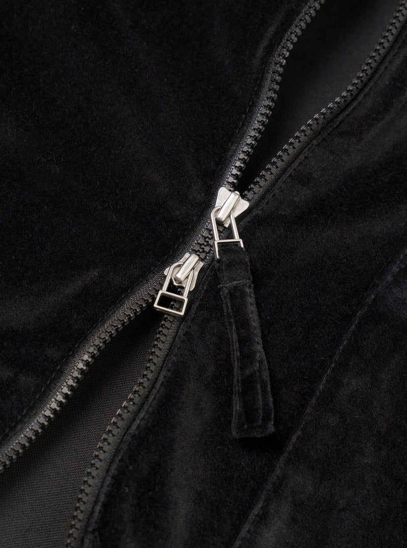 Close-up detail of the black velvet zip-front shirt, showing a two-way silver-toned zip with black plastic teeth, a black zip tape, and a fabric pull tab in matching black velvet. The soft, dense pile of the velvet fabric is visible on either side of the open zip.