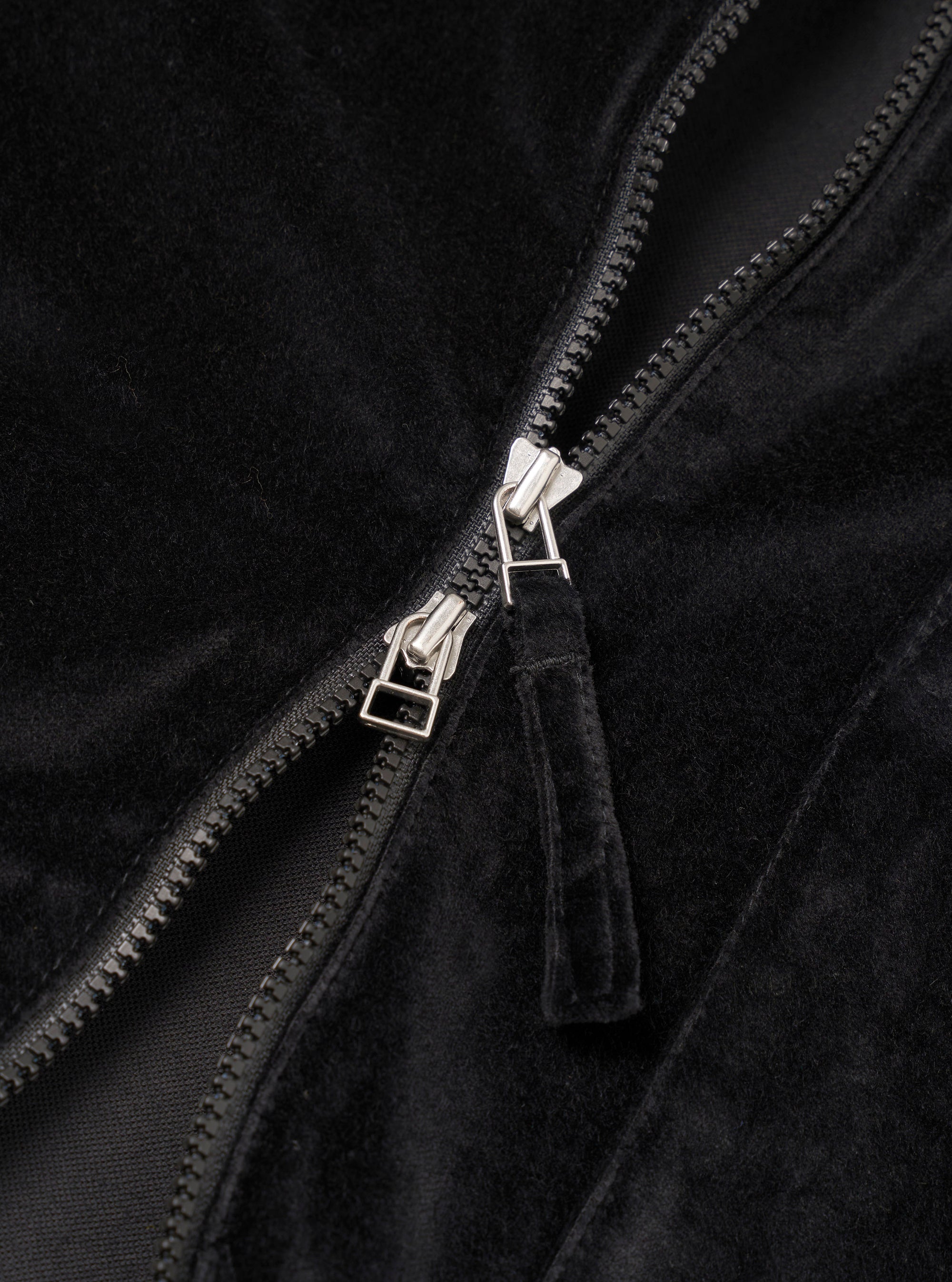 Close-up detail of the black velvet zip-front shirt, showing a two-way silver-toned zip with black plastic teeth, a black zip tape, and a fabric pull tab in matching black velvet. The soft, dense pile of the velvet fabric is visible on either side of the open zip.