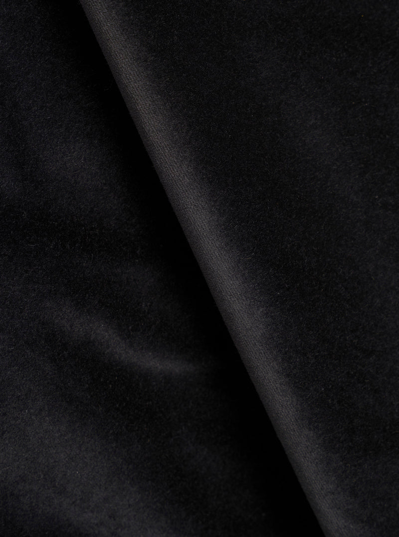 Close-up detail shot of black velvet fabric, showing a soft, dense pile texture with a diagonal fold running from top to bottom. The surface catches light subtly, highlighting the plush nap of the velvet. No garment construction details are visible.
