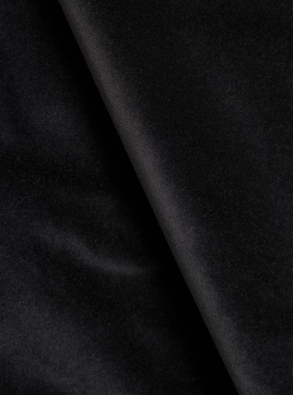 Close-up detail shot of black velvet fabric, showing a soft, dense pile texture with a diagonal fold running from top to bottom. The surface catches light subtly, highlighting the plush nap of the velvet. No garment construction details are visible.