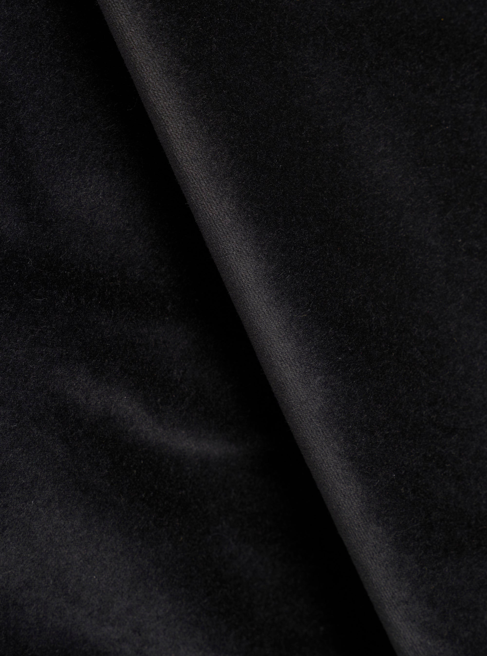 Close-up detail shot of black velvet fabric, showing a soft, dense pile texture with a diagonal fold running from top to bottom. The surface catches light subtly, highlighting the plush nap of the velvet. No garment construction details are visible.