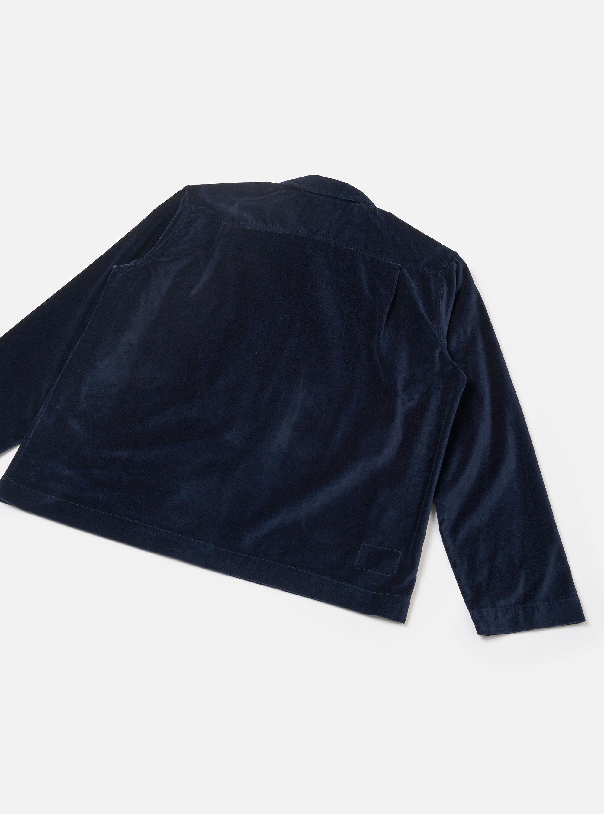 Navy velvet zip shirt laid flat on a light grey surface, shot from the back at an angle. The smooth, lustrous velvet fabric has a fine corduroy-like texture. Visible details include a back yoke seam, a straight cropped hem with topstitching, long sleeves with plain cuffs, and a small tab detail at the lower back hem.
