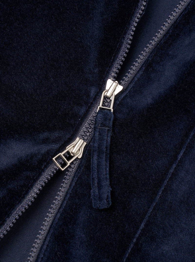 Close-up detail of the navy velvet zip-front shirt. A two-way silver-tone zip runs diagonally across the frame, partially open, with a fabric-covered navy velvet zip garage visible. The dense, soft velvet pile and a smooth navy lining are visible either side of the zip teeth. Plain white studio backdrop.