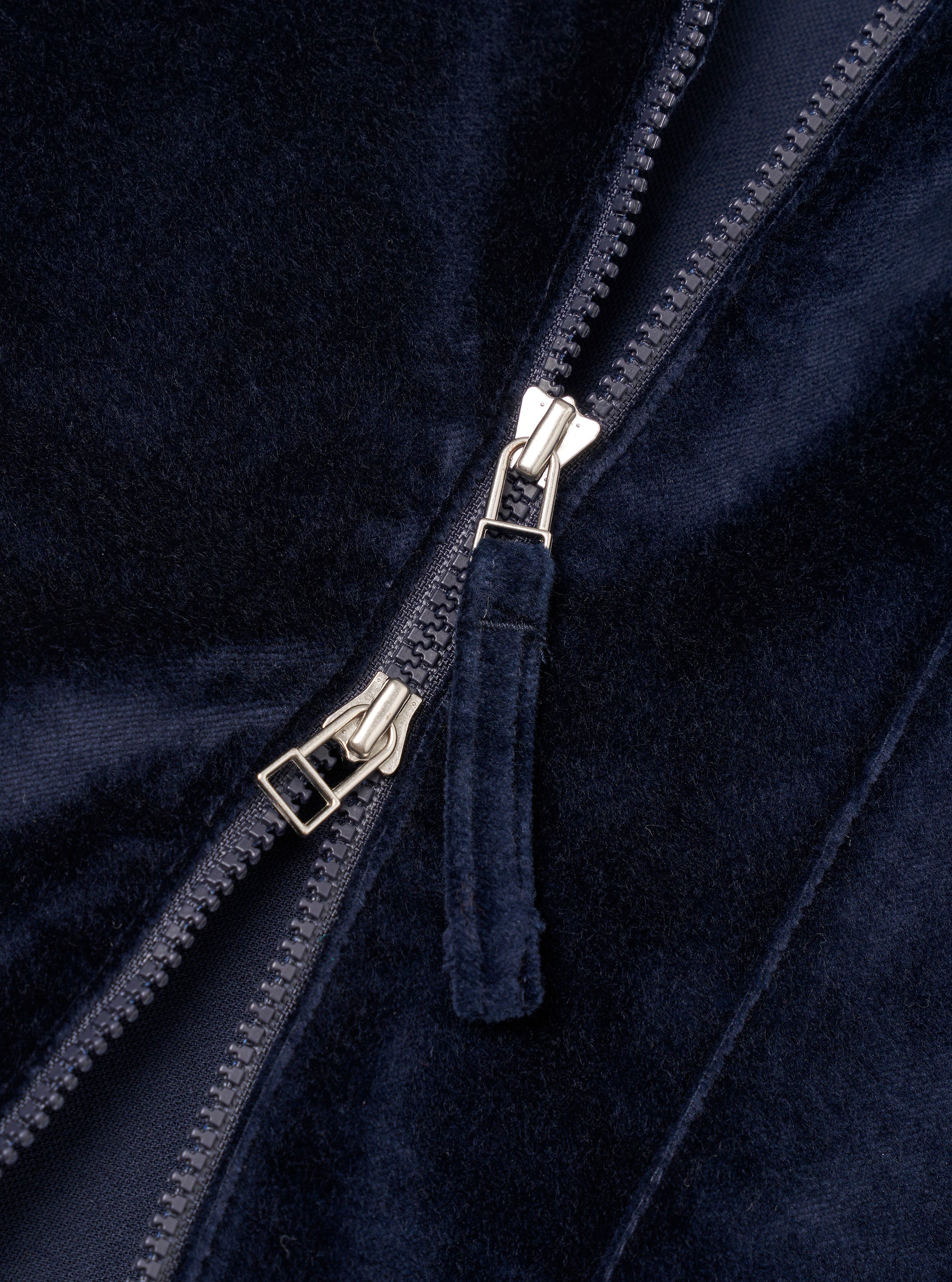 Close-up detail of the navy velvet zip-front shirt. A two-way silver-tone zip runs diagonally across the frame, partially open, with a fabric-covered navy velvet zip garage visible. The dense, soft velvet pile and a smooth navy lining are visible either side of the zip teeth. Plain white studio backdrop.