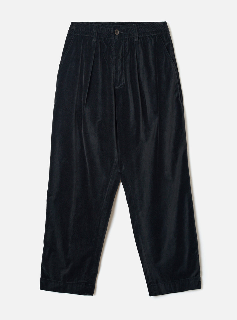 Black velvet Oxford trousers laid flat on a light grey seamless backdrop. The wide-leg, tapered silhouette features a double-pleated front, elasticated waistband with a single button fastening, and side seam pockets. The fine-wale velvet fabric has a subtle sheen. Hem is plain and unfinished at the ankle.