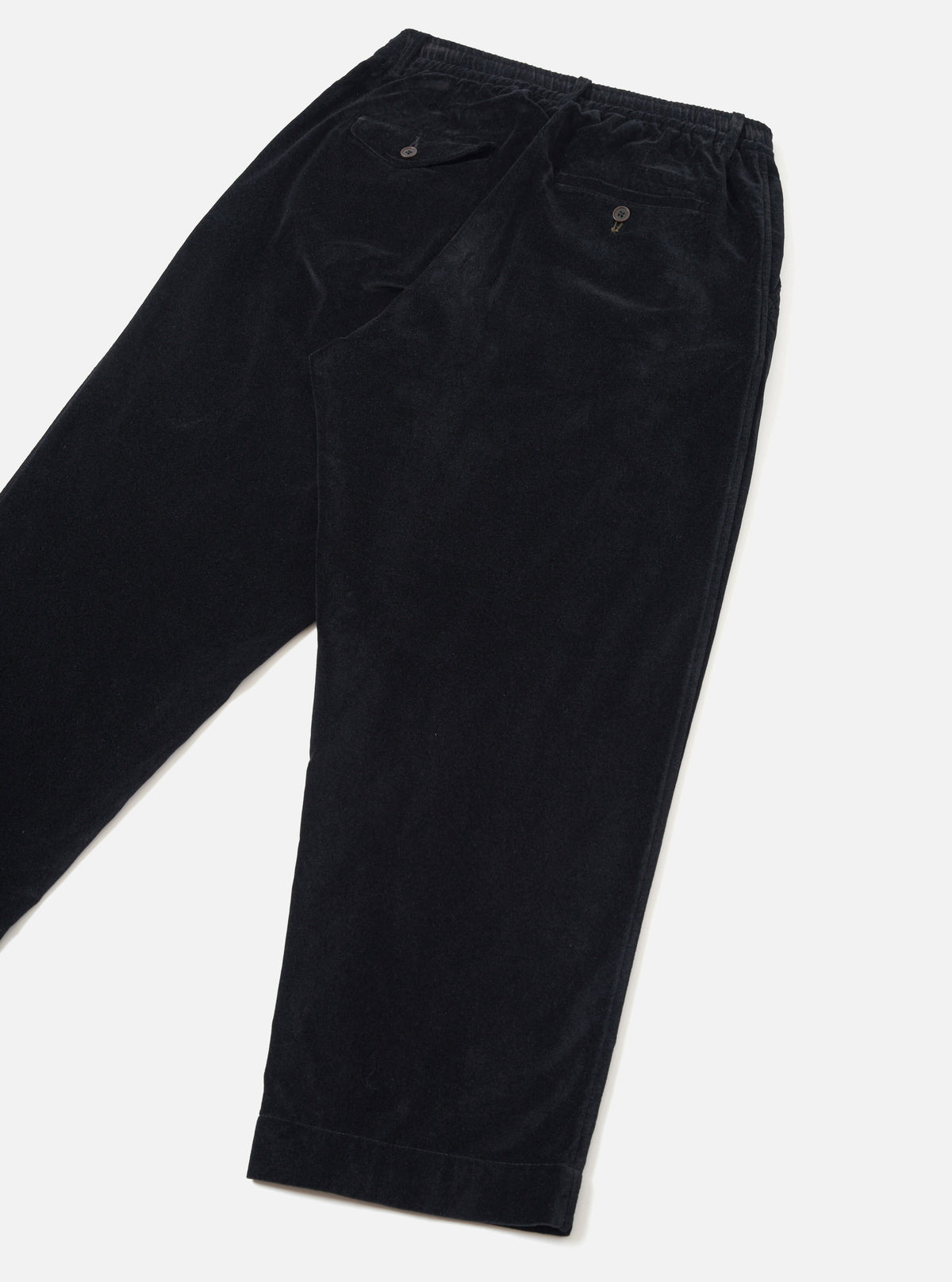 Black velvet corduroy Oxford trousers in a flat lay on a light grey seamless backdrop. The elasticated waistband, two buttoned rear welt pockets, and fine ribbed velvet texture are visible. The trousers are folded and cropped above the knee, showing a relaxed leg with plain, unfinished hems.