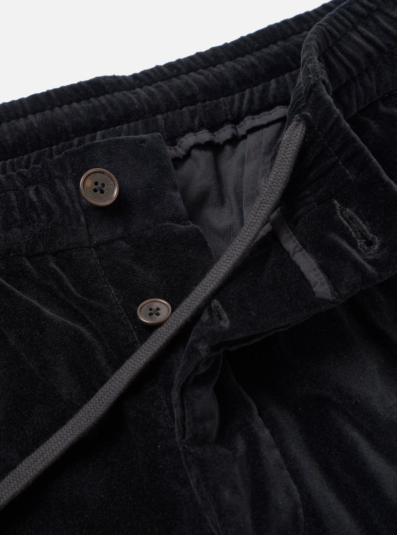 Close-up detail of black velvet Oxford trousers on a white studio backdrop. The elasticated corduroy-ribbed waistband sits at the top of the frame. The fly is open, revealing two four-hole horn-effect buttons on the button fly and a dark fabric lining. A grey woven drawcord runs diagonally across the front.