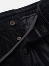 Close-up detail of black velvet Oxford trousers on a white studio backdrop. The elasticated corduroy-ribbed waistband sits at the top of the frame. The fly is open, revealing two four-hole horn-effect buttons on the button fly and a dark fabric lining. A grey woven drawcord runs diagonally across the front.
