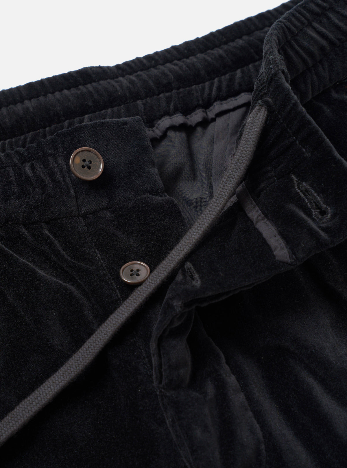 Close-up detail of black velvet Oxford trousers on a white studio backdrop. The elasticated corduroy-ribbed waistband sits at the top of the frame. The fly is open, revealing two four-hole horn-effect buttons on the button fly and a dark fabric lining. A grey woven drawcord runs diagonally across the front.