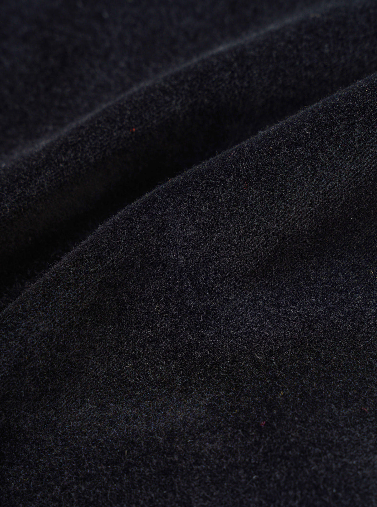 Close-up detail shot of the black velvet fabric used in the CPH Oxford Pant. The surface shows a dense, soft pile with a fine brushed texture and subtle dark navy undertones. Occasional flecks of red and orange fibre are visible throughout. The fabric is folded, highlighting its weight and drape.