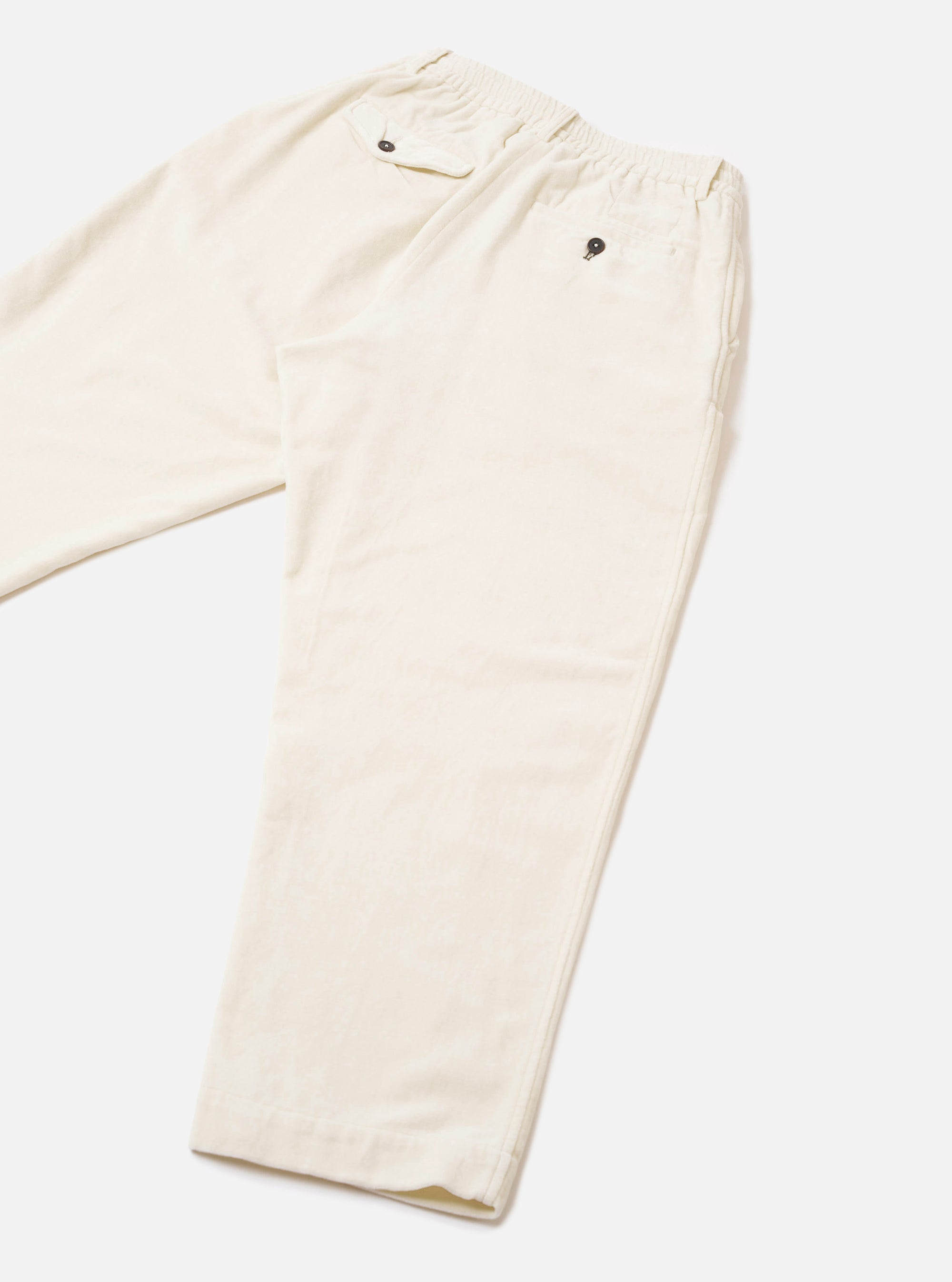 Ecru velvet Oxford trousers laid flat on a light grey seamless backdrop. The elasticated waistband features a small metal eyelet detail at the front. Fine velvet texture is visible across the fabric. Side seams run the full length of the leg to the plain hem.