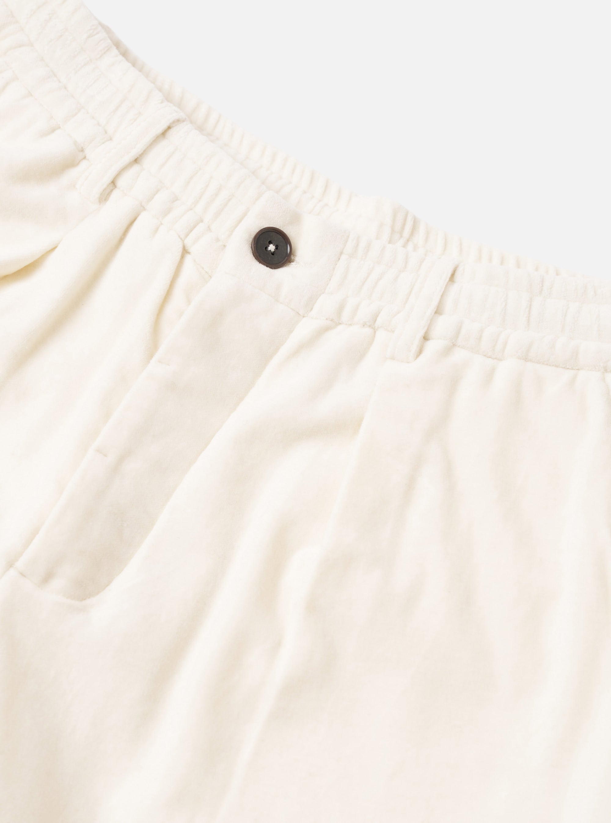 Close-up of ecru velvet Oxford trousers against a light grey studio backdrop. The ribbed elastic waistband is shown in detail alongside a single dark horn button at the fly. A slash pocket is visible to the left. The smooth velvet fabric has a soft, matte texture.