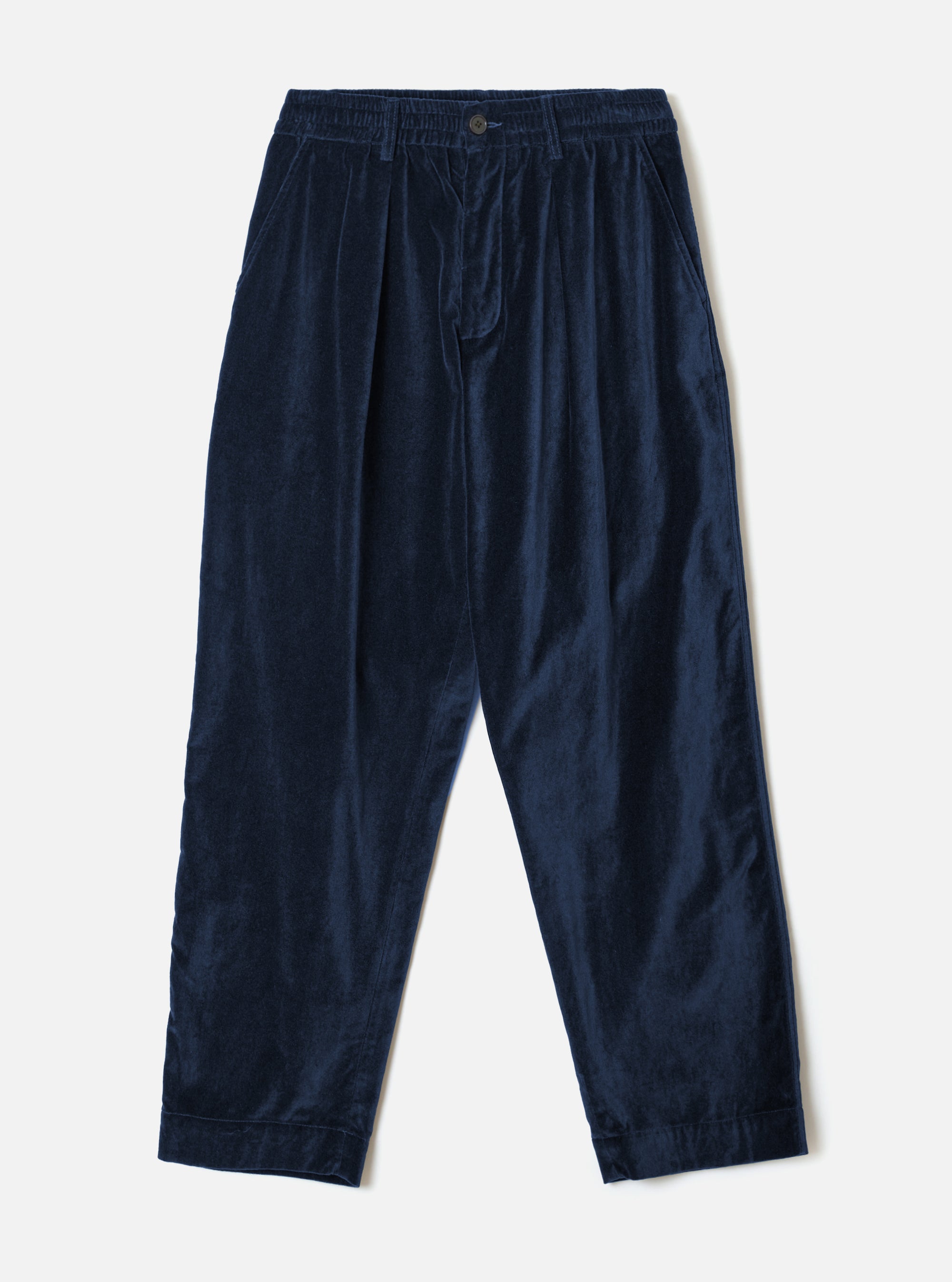 Navy velvet Oxford trousers laid flat on a light grey studio backdrop. Fine-wale velvet fabric with a subtle sheen. Elasticated waistband with a single centre button, double forward pleats, side slash pockets, and tapered legs with plain hems.